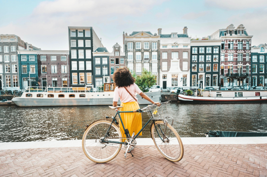 Frau mit gelben Rock und wei&szlig;em Shirt lehnt an ihrem Fahrrad mit Blick auf eine H&auml;userreihe an einer Gracht in Amsterdam